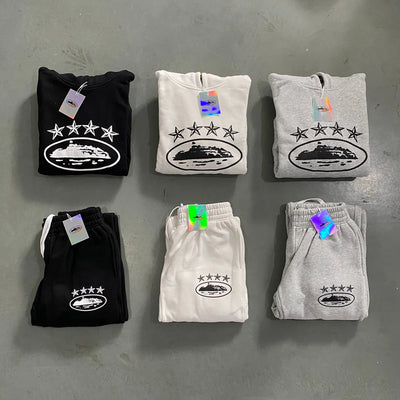 Folded Corteiz 4Starz Alcatraz Hoodie Tracksuits in black, white, and gray are elegantly arranged on the floor. Each streetwear piece boasts the signature logo with stars and a central design. Clothing tags hang from these premium cotton garments, some featuring iridescent colors.